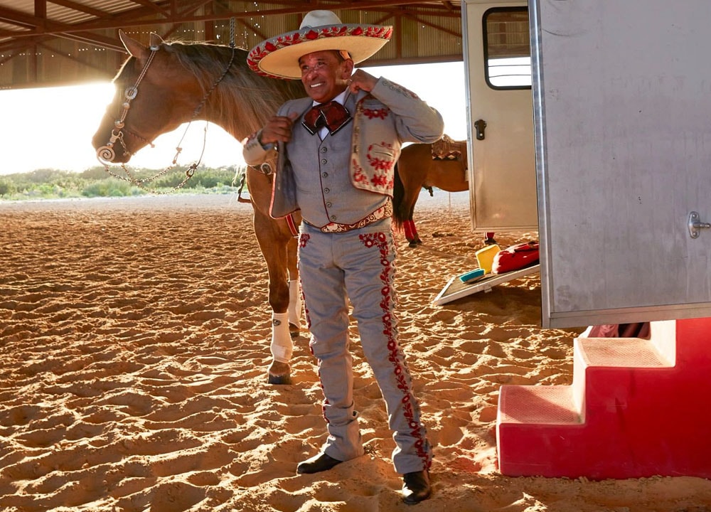 Un hombre vestido con sombrero y traje tradicional de Charro poniéndose una chaqueta frente a un caballo y de pie junto a un remolque para caballos.